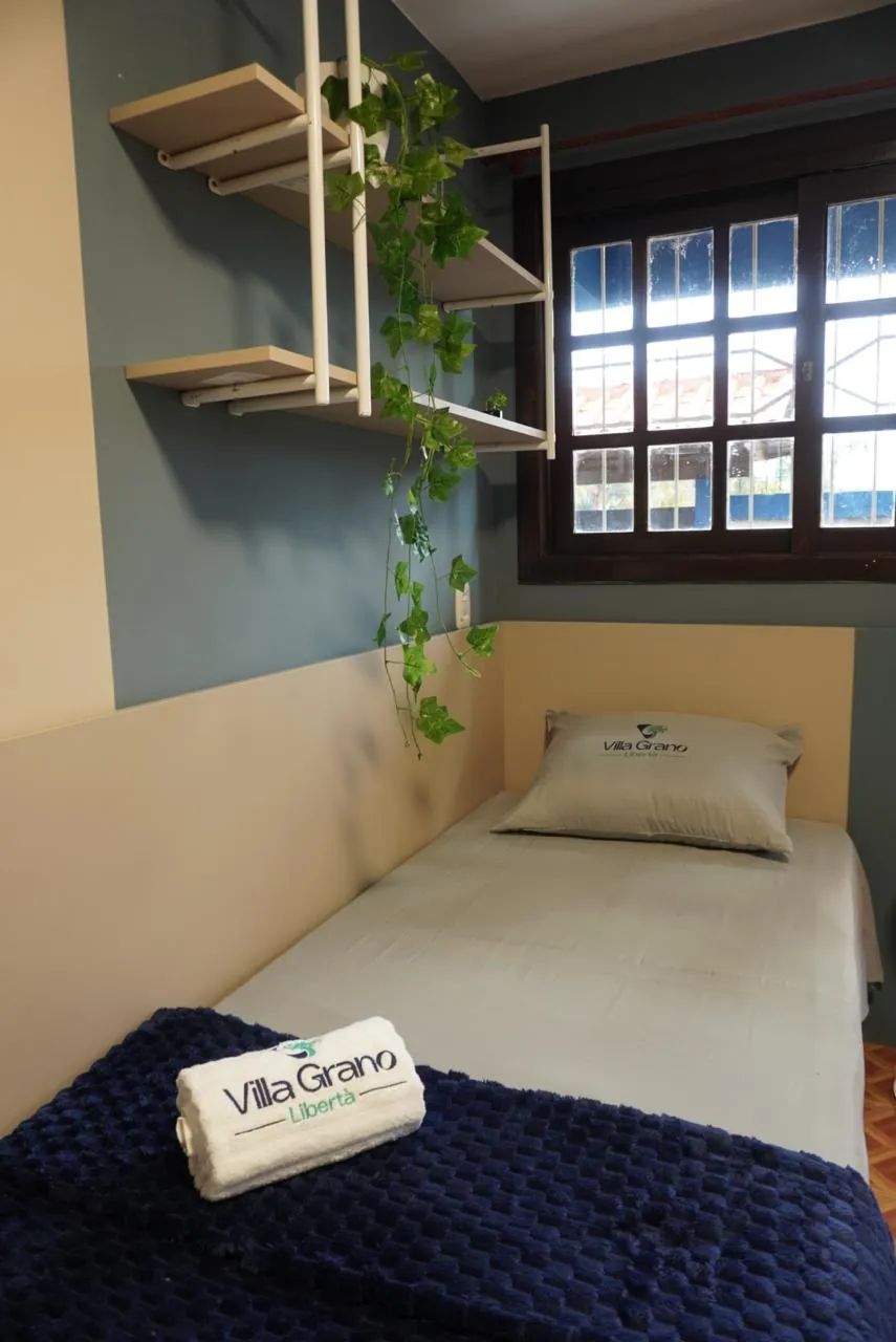 Quarto com cama de solteiro, janela de madeira e prateleiras com plantas
